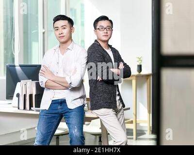 portrait of two young asian business men posing back to back in office looking at camera Stock Photo