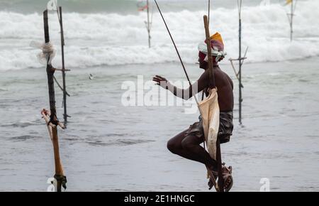 Weligama, Sri Lanka - 07 26 2020:Old Fisherman holding a rode in a wooden pole while sitting, fishing in the evening, catch a small fish, a reward for Stock Photo