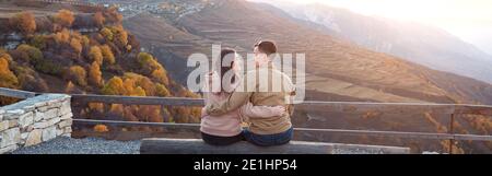 Romantic couple in love kisses and hugs sitting on large log of observation deck against sunrise over rocky hills and orange forests backside view Stock Photo