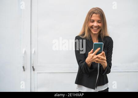 Outdoor portrait of happy modern businesswoman. Blonde businesswoman is standing outdoor and looking at smartphone. Stock Photo