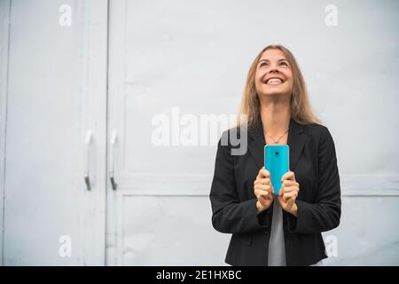 Outdoor portrait of happy modern businesswoman. Blonde businesswoman is standing outdoor and looking at smartphone. Stock Photo