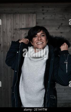 Cheerful woman wearing winter clothes putting her hood on while smiling at camera with a perfect smile Stock Photo