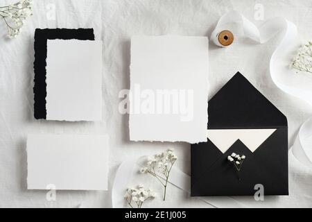 Elegant set of wedding stationery top view. Flat lay blank paper cards, black envelopes, flowers, ribbon on white textile background. Stock Photo