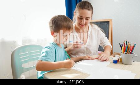 Smiling mother looking at her little son drawing or writing while sitting behind desk at home classroom. Concept parents and children of education and Stock Photo