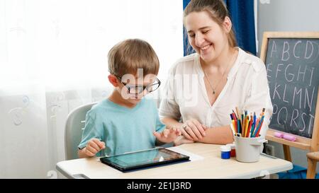 Little boy in eyeglasses playing on tablet computer. Mother hugging her little son doing homework using gadet. Children having problems with eyes and Stock Photo