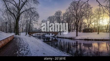 Panoramic winter landscape with sunrise in snowy park with beautiful bridge over small canal, street light and covered in snow tress. View of Bastion