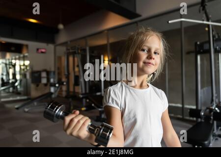 Little cute adorable caucasian blond girl doing finess exercise lifting dumbbell in sport gym club indoors. Happy sporty tired child. Children Stock Photo