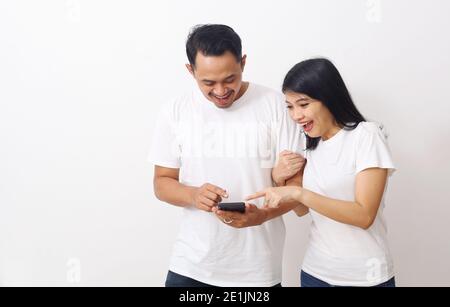 Happy asian couple surprised while looking and pointing their smartphone. Get good news concept from smartphone. Isolated on white with copyspace Stock Photo