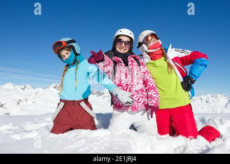 Three teenage girls on winter hollidays in mountain sitting in fresh snow look at the camera. Landscape with big blue sky and colorful clothes Stock Photo