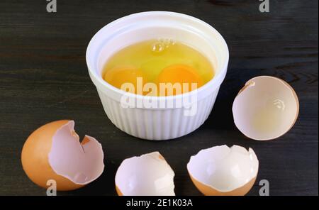 Closeup two of raw eggs in a bowl on black wooden table with cracked eggshells in foreground Stock Photo