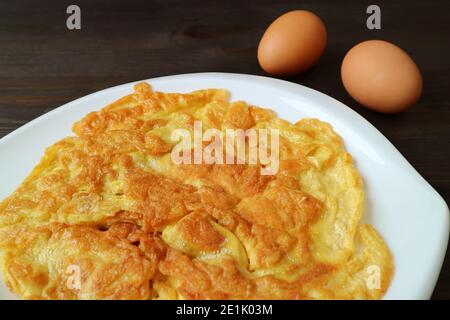 Plate of Thai Style Omelette with Two Raw Eggs on Dark Brown Wooden Table Stock Photo