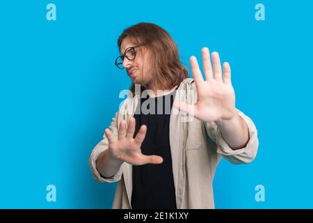 Confident man with long hair and beard is gesturing the refuse sign with palms on a blue studio wall Stock Photo