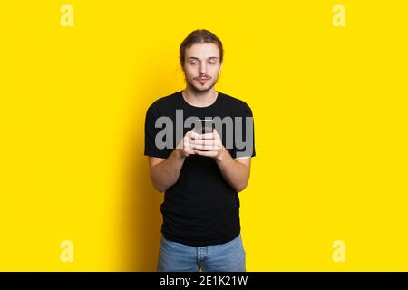 Caucasian man with beard and long hair is chatting on mobile wearing a black t-shirt on a yellow studio wall Stock Photo