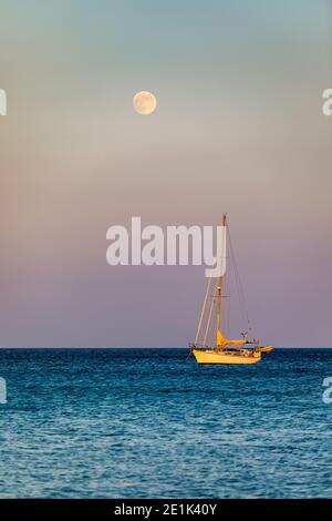 Full moon rising over the water with a small sailing boat in the foreground. Sailing boat with raising moon at sunset. Moon rising over the sea and ya Stock Photo