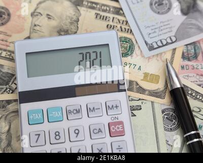2021 is shown on a calculator LCD screen among multiple denominations of USA dollar bills and next to an ink pen. Stock Photo