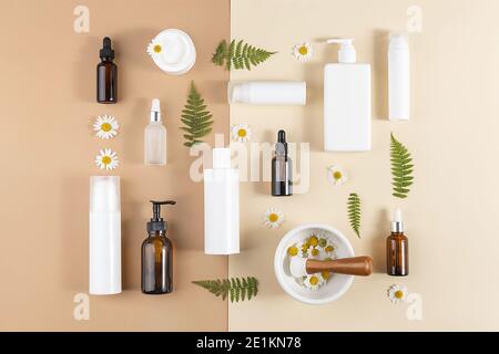 Set of care cosmetics. Various bottles, tubes with cosmetic, chamomile flowers, fern leaves and mortar bowl with pestle on beige and brown background. Stock Photo