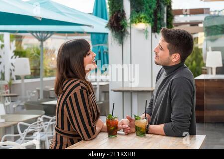 view of a young couple enjoying a talk and cocktails at an outdoor bar terrace on an out of focus background. Couples and leisure concept. Stock Photo