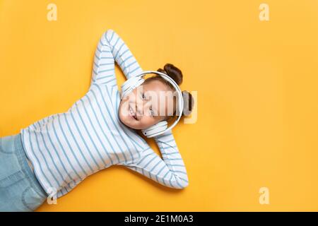 Cheerful cute little girl in a good mood listening to modern technology headphones on a yellow background. The child lies with his hands behind his he Stock Photo