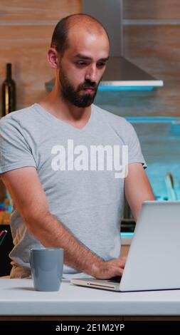 Tired sleepy man yawning, working at home desk in the kitchen. Busy overwork focused employee using modern technology network wireless doing overtime for job reading writing, searching Stock Photo