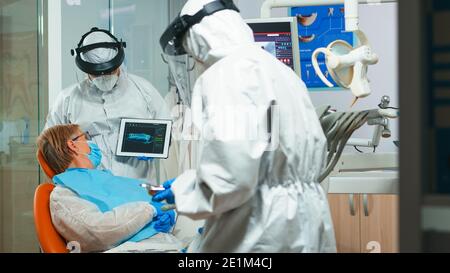 Dentist in coverall using tablet explaining dental x ray to patient in stomatologic office during coronavirus. Man wearing face shield, coverall, mask showing to woman radiography using digital device Stock Photo