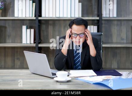 Sad Asian businessman holds his temple with both hands, sitting in the office. The concept of unemployed, sadness, depressed and human problems. Stock Photo
