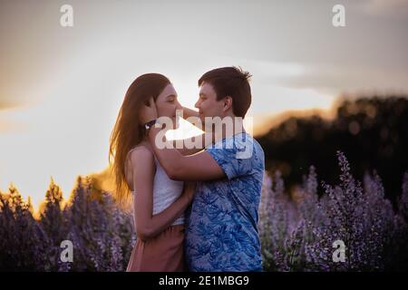 Happy couple in love hugs, kisses at sunset near a blooming sage field. A young man passionately looks into the eyes, holding a beautiful girl by the Stock Photo