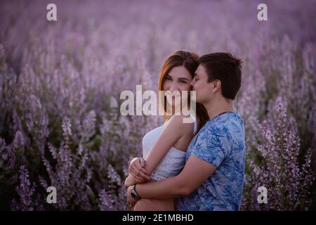 Happy couple in love hugs, kisses at sunset near a blooming sage field. A young man passionately looks into the eyes, holding a beautiful girl by the Stock Photo