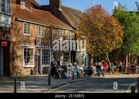 Tables & chairs outside quaint street cafes, people sitting in autumn sun, relaxing, having refreshments - scenic College Street, York, England, UK Stock Photo