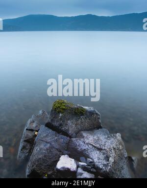 Moody and peaceful early morning long-exposure shot of a moss-covered stone on the shore of Ashinoko Lake in the mountains of Hakone, Japan. Stock Photo