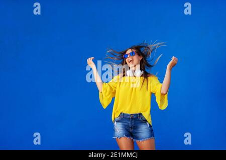 Cheerful girl in yellow shirt standing against blue wall with headset around her neck. Dancing and smiling. Stock Photo