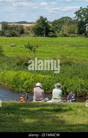 Elderly hiking couple taking a picnic break on the river bank and enjoying nature. Rewe village, Devon, England. Stock Photo