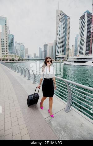 Busy young modern business woman in suit pulling a suitcase in a Dubai Marine. Walking to her office. Stock Photo