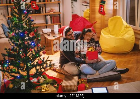 Man holding gift in one hand and with other covering woman's eyes. Couple sitting on the floor. Next to them Christmas tree. Christmas holidays concep Stock Photo