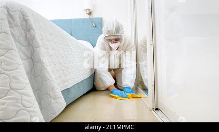 Housekeeper or maid in hotel wearing protective medical suit and mask washing and cleaning floor in room. Disinfection in hotels Stock Photo