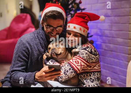 Happy couple reading text message on smart phone while sitting on floor. GIrl holding dog. On heads santa's hat. Christmas holidays concept. Stock Photo