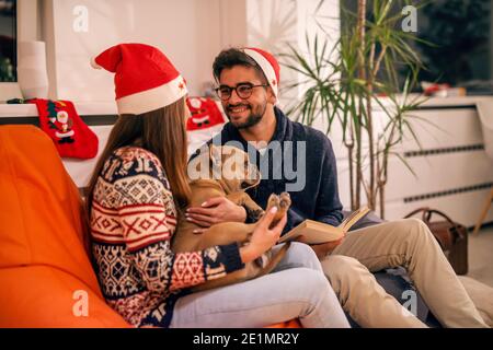 Couple spending Christmas eve at home. Girl holding dog and man reading a book. On heads santa's hat. Christmas holidays concept. Stock Photo