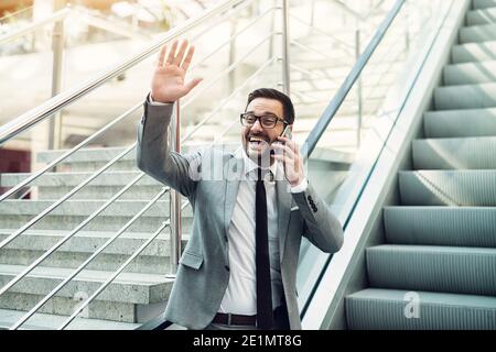 Cheerful modern busy businessman talking over cell phone and waving to his friends. Stock Photo