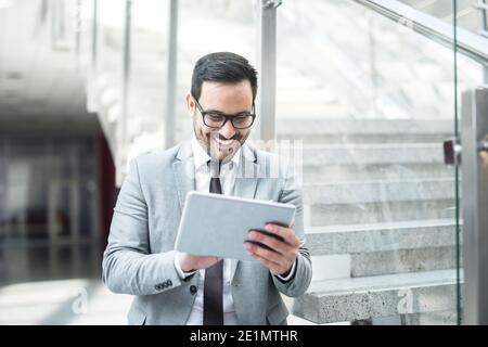 Happy young Caucasian businessman using tablet for checking mail while standing in the hall. Stock Photo