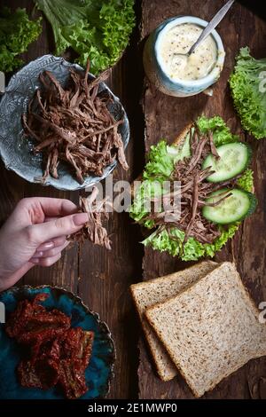 hands preparing meat sandwich with green lettuce leaves, dry tomatoes, fresh cucumbers and rye or wheat bread, on wooden dark rustic table background, Stock Photo