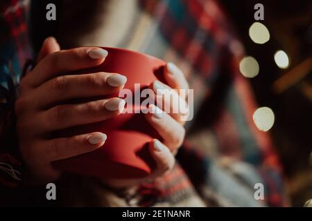Unrecognizable woman holding cozy red mug on glowing Christmas tree background. Girl with hot drink - tea, coffee or cocoa. Concept of the New Year Stock Photo