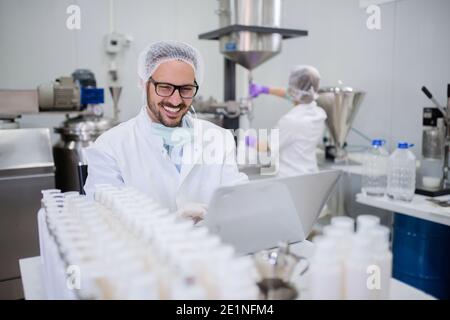 Technologist using laptop for entering test results while sitting in the lab. In background his assistant testing cosmetic products. Stock Photo