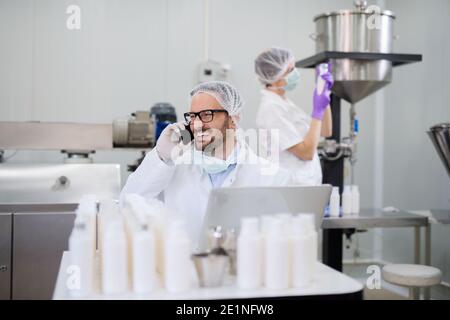 Man in working suit talking on the mobile while sitting in the lab. In background his assistant holding cosmetic product. Stock Photo