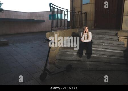 Young woman taking with the cell phone sit-in on the stairs with a e-scooter Stock Photo