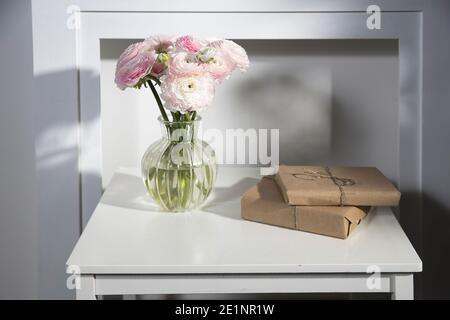 A bouquet of pink Persian buttercups in a glass vase and two wrapped gifts on a white table in front of a fake white fireplace. Stock Photo