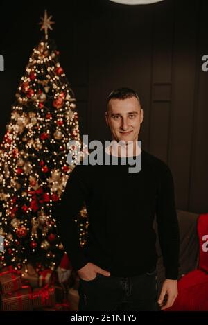 handsome young man in black clothes near a Christmas tree in garlands. decorated house for New Year. Christmas morning. apartment interior. Valentine' Stock Photo
