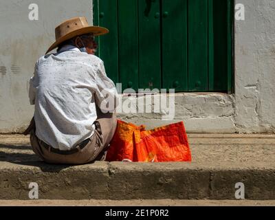 A man with a hat sitting on a sidewalk in front of a green door in a street of the colonial town of Villa de Leyva, in the central Andean mountains of Stock Photo