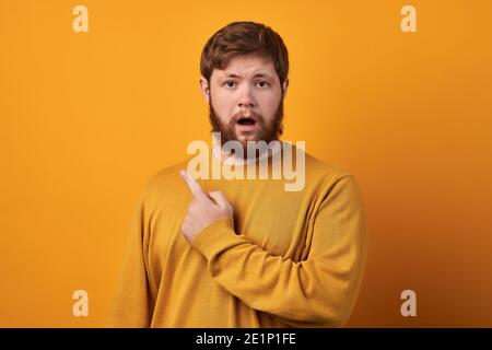 Close up shot of emotive shocked bearded young male opens mouth widely, stare at camera, points upwards aside at blank space, says: Wow, it s somethin Stock Photo