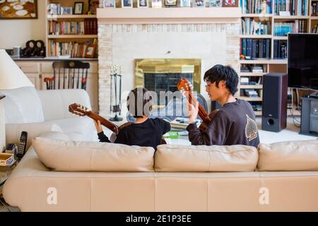 A father and son sit together on couch playing classical guitar music Stock Photo