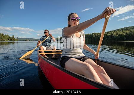 white woman and black man paddle red canoe on woodland lake in Maine Stock Photo