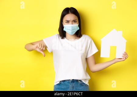 Covid-19 and real estate concept. Disappointed asian woman in medical mask, showing thumb down and paper house cutout, standing upset against yellow Stock Photo